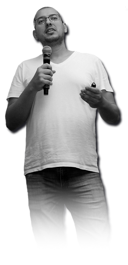 Foto de corpo de Marcelo Sales. A imagem está em preto e branco e Marcelo está em posição de apresentação com um microfone na mão direita e um passador de slides na mão esquerda. Está de camiseta branca e caça jeans.